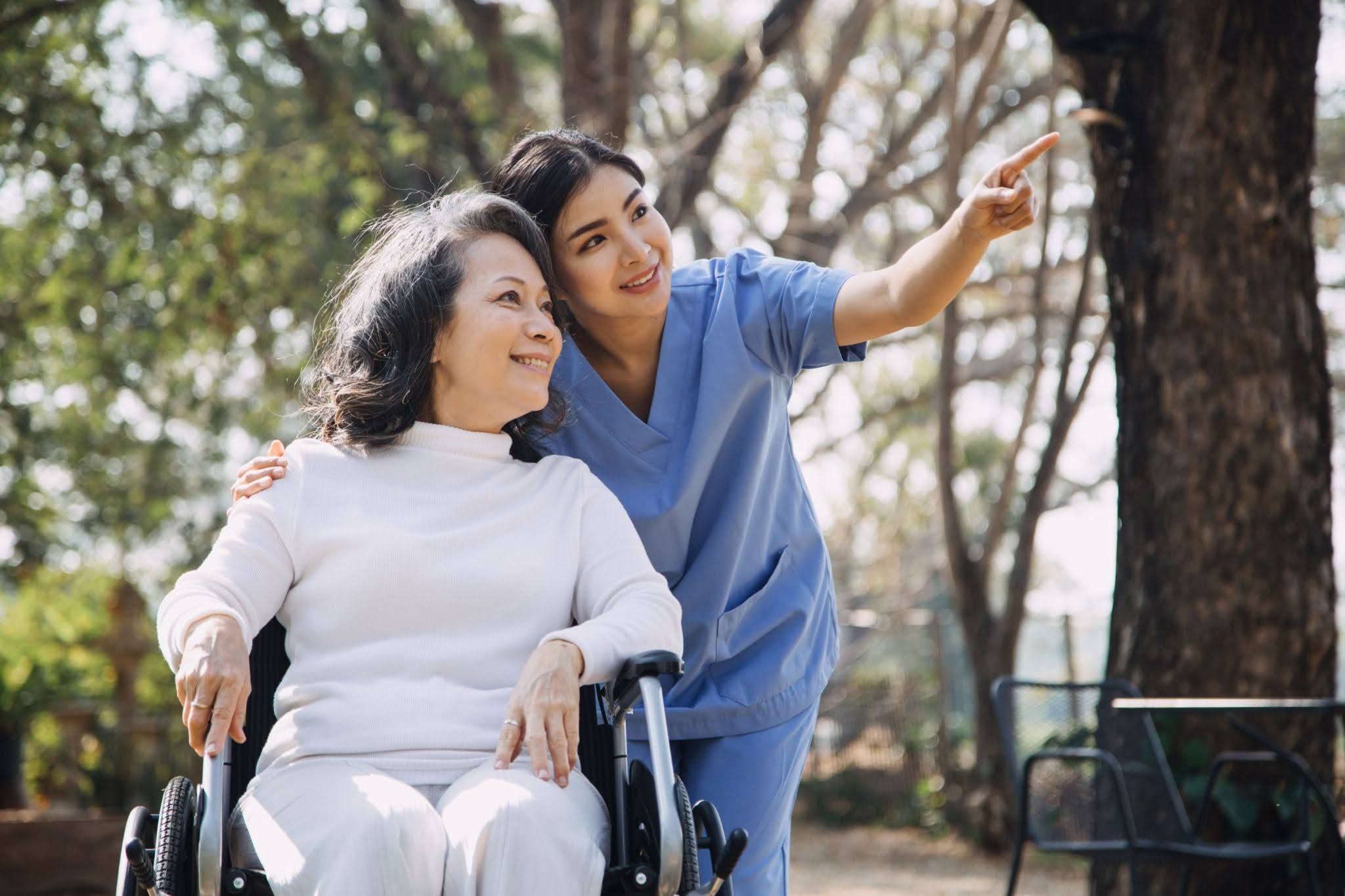A slightly older woman sitting in a wheelchair with a young caretaker woman hugging to her and pointing to something at a park